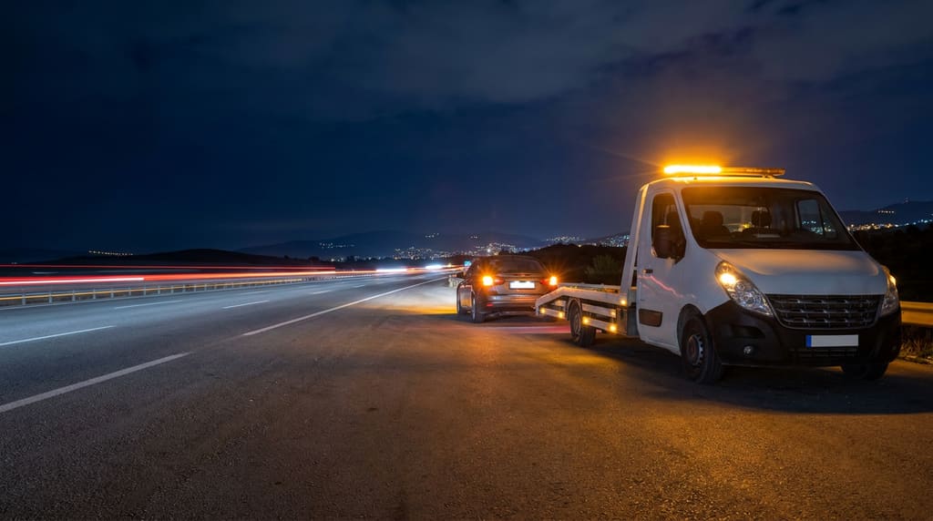 Gece otoyol kenarında turuncu tepe lambaları yanan beyaz bir kayar kasa çekici ve arkasında dörtlüleri yanan bir otomobil